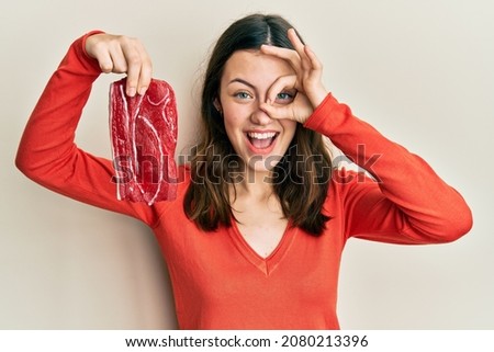 Young brunette woman holding raw beef steak smiling happy doing ok sign with hand on eye looking through fingers 