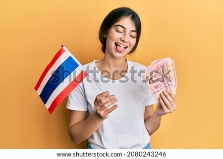 Young caucasian woman holding thailand flag and baht banknotes sticking tongue out happy with funny expression. 