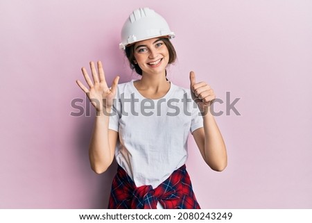 Young caucasian woman wearing hardhat showing and pointing up with fingers number six while smiling confident and happy. 