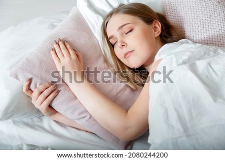 Teenager girl in pajamas sleeps in bedroom at morning time. Young Woman sleeping in bed. Portrait of blonde teen girl have healthy good sleep on white pink pillow.