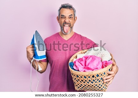 Handsome middle age man with grey hair holding electric steam iron holding laundry basket sticking tongue out happy with funny expression. 