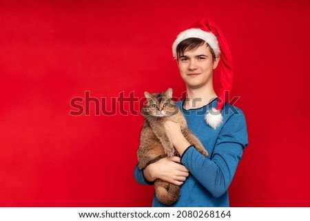 Young man in Santa's cap holds a cute British cat on a red background. Copy space.