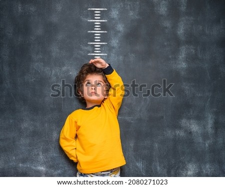 Funny little kid boy measuring his growth in height. Child in yellow clothes looking up with hand above his head. Time to grow. Kindergarten kid is ready to go to school.
