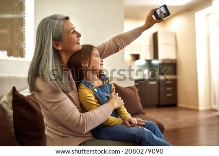 Portrait of happy mature woman and cute young girl sitting on sofa, taking selfie. Happy moments with nanny or babysitter. Taking photo to remember good moments spent together.