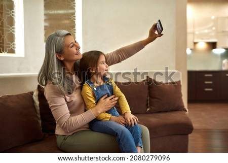 Side view cheerful grey-hair senior woman sitting on couch, holding cute little girl on her laps, holding smartphone. Smiling granny and grandaughter taking selfie at home.