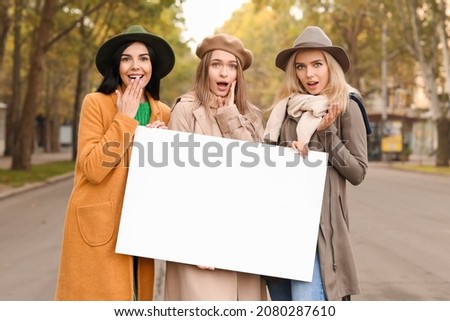 Beautiful surprised young women with blank poster outdoors