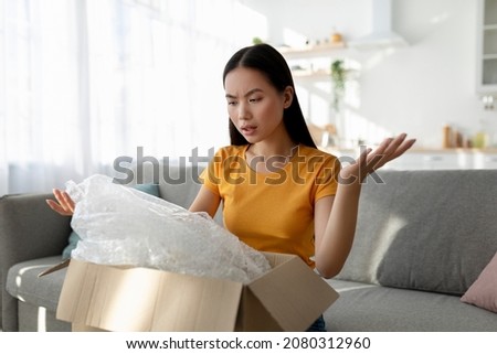 Terrible delivery service. Unhappy asian woman unboxing cardboard parcel, receiving damaged item, sitting on sofa at home. Young lady feeling disappointed about wrong parcel Royalty-Free Stock Photo #2080312960