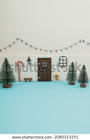 Mini elf or mouse house diy toy gift for kids made with tiny door, window with lantern and Christmas green tree of pipe brushes. Little bulb garland on white wall. Holiday decor on blue background.