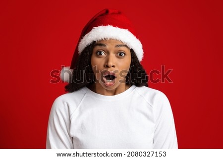 Closeup portrait of amazed attractive young black lady in santa hat looking at camera with eyes and mouth wide open, red studio background. New Year 2022, Christmas celebration concept