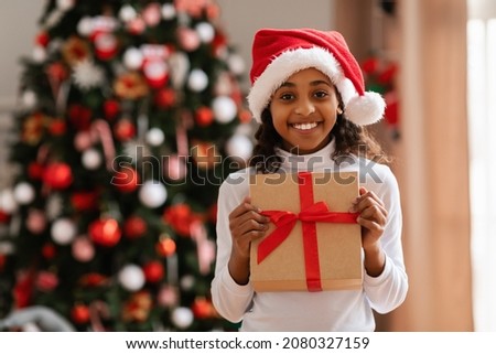 Holidays And Celebration Time. Portrait of excited beautiful black girl in red Santa Claus hat holding present box posing in decorated living room. Smiling teen showing gift wrapped in craft paper