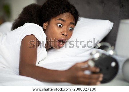 Shocked african american young woman laying in bed and looking at alarm on bedside table in bedroom at home, overslept. Black lady is late for appointment or work or class study, copy space Royalty-Free Stock Photo #2080327180