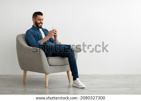 Joyful young Arab guy sitting in armchair with smartphone, working remotely or video chatting against white studio wall, empty space. Middle Eastern man watching movie, having online meeting Royalty-Free Stock Photo #2080327300