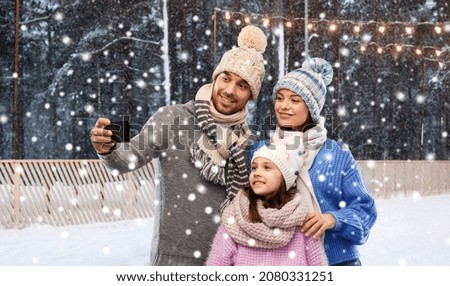 family, christmas and winter holidays concept - happy mother, father and little daughter in winter clothes taking selfie by smartphone over ice rink background