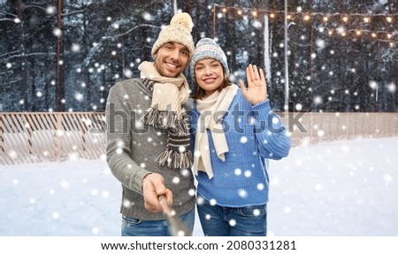 people, christmas and winter holidays concept - happy couple in knitted hats and scarves taking picture by selfie stick over ice rink background