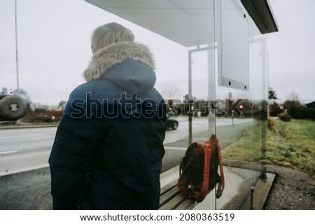 Man in blue coat with backpack is waiting for a bus. Modern glass bus stop on Norway. Cloudy day.