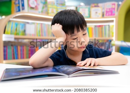Adorable young Asia boy put his hand on the head and sitting on the chair in the modern library or bookstore and reading a book. Education, learning concept. Kid interest cartoon book. Feeling bored