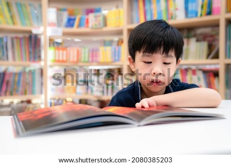 Adorable young Asia boy wearing blue shirt and sitting on the chair in the modern library or bookstore and reading a book. Education and learning concept. Kid interest cartoon book. Feeling happy