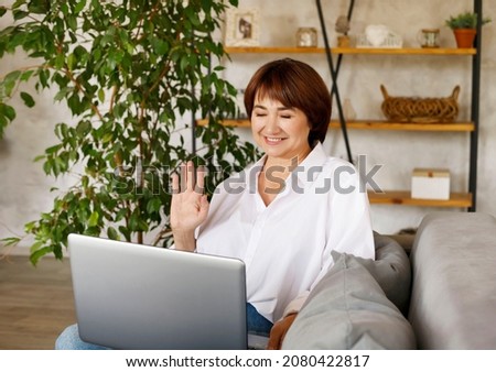 Middle aged woman working or learning on her laptop showing hand in notebook sitting in living room in quarantine lockdown. Social distancing communication.