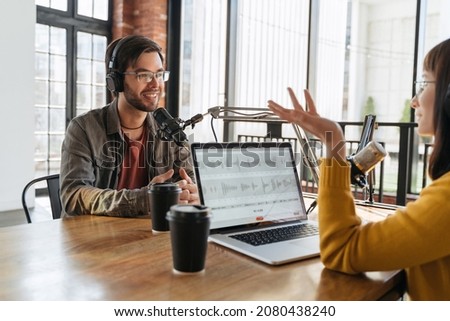 Cheerful young man blogger talking with gesturing woman radio host, smiling, have an interview for audio podcast in studio. Young man and woman recording live stream. Social media, content creators
