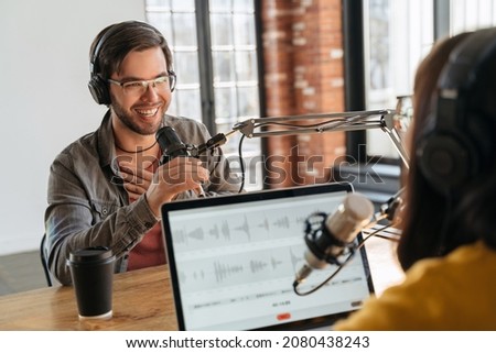 Podcast content. Excited handsome male guest gesturing, looking at female host while they making audio podcast in studio. Two podcasters in headphones laughing, talking, recording live podcast Royalty-Free Stock Photo #2080438243
