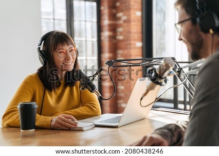 Young beautiful woman radio hosts smiling while interviewing man guest in studio while recording podcast for online show. Two podcasters in headphones looking at each other and recording audio podcast