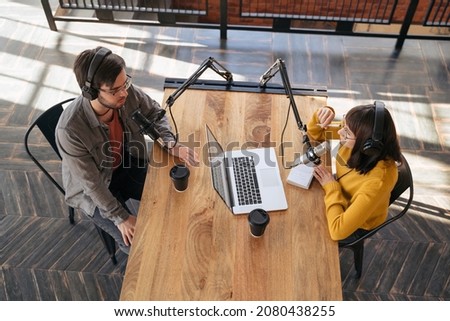 Two young radio hosts in headphones are talking while sitting near microphones and making audio podcast in broadcasting studio. Top view. Social media, podcast creating, content creator, blogging