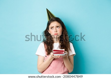 Holidays and celebration. Sad girl in party hat holding birthday cake, looking away with thoughtful upset grimace, feeling lonely and moody on her bday, standing over blue background