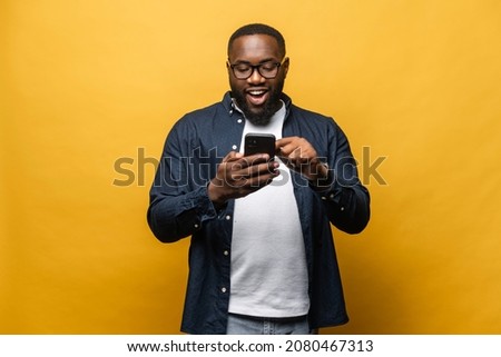 Portrait of cheerful young black guy sending message on mobile phone against yellow background. Happy African-American man using smartphone, enjoying chatting online, messaging in social networks