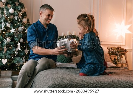 A young father gives Christmas gifts to his smiling daughters near a decorated Christmas tree.