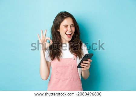 Online shopping. Cheerful cute girl winking at you, smiling and showing okay sign after using smartphone app, recommending internet shop or social media page, blue background