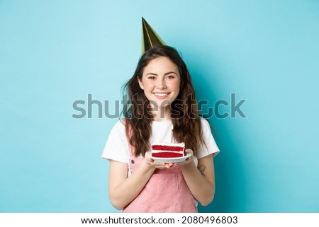 Holidays and celebration. Cheerful birthday girl in party hat holding bday cake and smiling, making wish on lit candle, standing against blue background