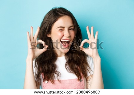 Close up of cheerful caucasian woman agree and say yes, make alright okay sign and winking assuringly at camera, standing satisfied over blue background
