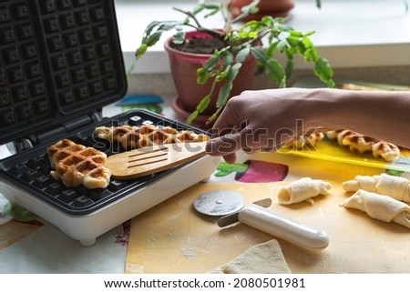 A young man removes a baked waffle from a waffle iron with a wooden spatula. Next to it are blanks of raw, puff-yeast dough, rolled into rolls. Asian dessert, culinary trend..