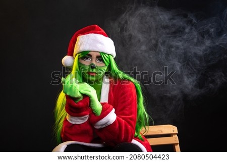 woman disguised as christmas, closeup portrait of mean, grumpy, unhappy, annoyed funny looking young woman, sitting in studio black background with santa claus costume green wig lady greem with smoke