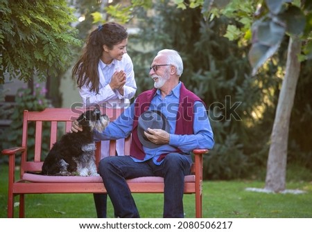 Elderly man with dog sitting in park while pretty nurse in white coat taking care of patient
