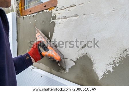 Construction worker covering house wall with adhesive cement glue berore installing styrofoam insulation sheets for thermal protection. Royalty-Free Stock Photo #2080510549