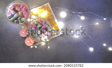 Colored marshmallows in a transparent glass for cocoa and a Christmas garland. Top view of a Christmas decoration. Waiting for Santa Claus, a treat, Candles on garland lights