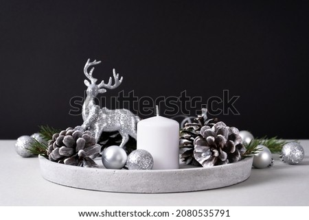 White candle on a concrete tray with pine cones, fir branches and silver Christmas balls with a deer on gray and black background. Interior for Christmas.