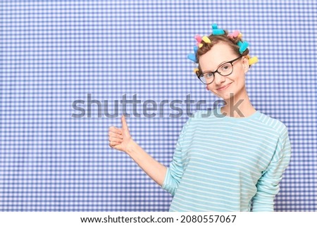 Portrait of happy young woman without makeup, with bright colorful hair curlers on head,  with glasses, smiling and raising thumb up in approval and like gesture, over shower curtain background
