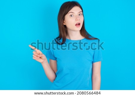 Emotive Young caucasian girl wearing blue T-shirt isolated over blue background keeps jaw dropped from shock demonstrates amazing promo points right on blank space demonstrates big shopping sale. 