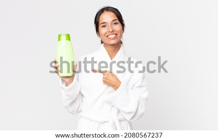 young hispanic woman smiling cheerfully, feeling happy and pointing to the side wearing bathrobe and holding a shampoo