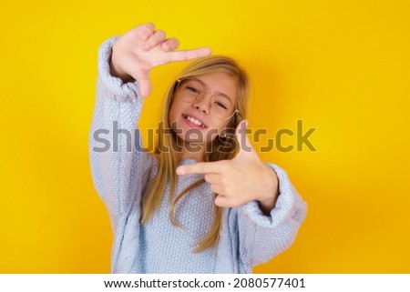 caucasian kid girl wearing blue knitted sweater over yellow background making finger frame with hands. Creativity and photography concept.