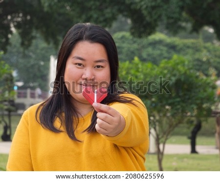 Portrait  girl Asian woman one people long black hair wearing yellow long-sleeved t-shirt. Sitting on park garden chair smile hand holding recycled paper happily folded into "gift heart" evening time