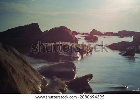 Close-up of rocks in seawater blurred by long exposure. Blue sky with clouds in the background. Beautiful red glare from the lens. Evening seascape Royalty-Free Stock Photo #2080694395
