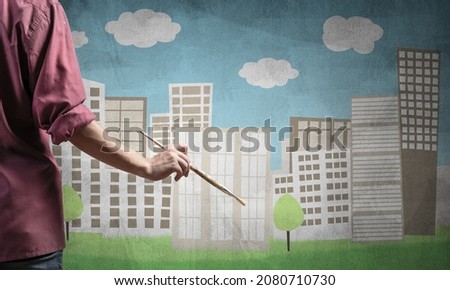 Close up artist hand holding paintbrush. Painter in shirt standing on background colorful picture of city. Modern cityscape with high skyscrapers artwork. Creative hobby and artistic occupation.