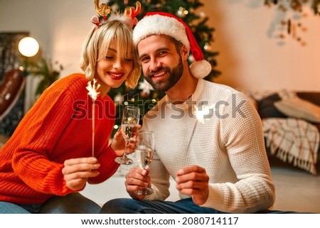 Merry Christmas and Happy New Year! A loving couple in Santa hats and reindeer antlers sitting on the floor near a Christmas tree with sparklers and glasses of champagne, waiting for a holiday.