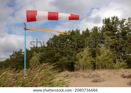 Windsock on a sandy beach in strong wind, Red and white fabric cone designed to indicate the direction and approximate wind speed, coastline, dense green forest Royalty-Free Stock Photo #2080736686