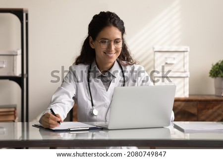 Happy young female GP doctor using laptop, making video call, reading electronic medical records. Medical practitioner giving online virtual consultation to patient from hospital office Royalty-Free Stock Photo #2080749847