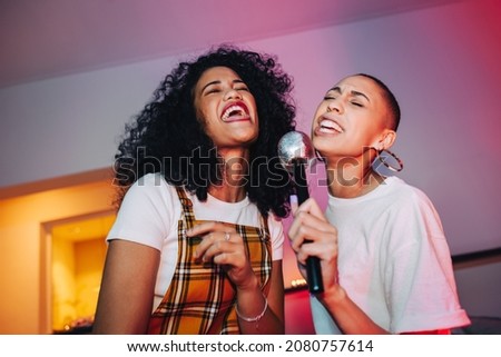 Best friends singing into a microphone on karaoke night. Two cheerful young women singing their favourite song at a house party. Happy female friends having a good time during the weekend. Royalty-Free Stock Photo #2080757614