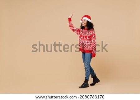 Full size body length friendly young african american Santa woman in Christmas hat sweater walk move go meet greet waving hand as notices someone isolated on pastel beige background studio portrait
